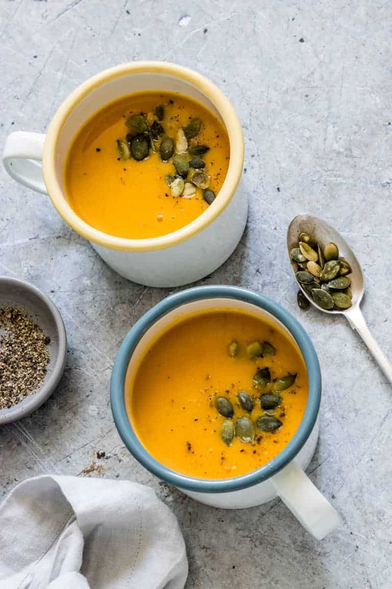 2 cups of vegan pumpkin soup on a table with pepper and pumpkin seeds