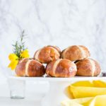Hot Cross Buns Recipe - Hot cross buns on a cake tray with flowers and a yellow cloth.