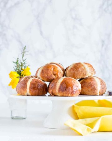 Hot Cross Buns Recipe - Hot cross buns on a cake tray with flowers and a yellow cloth.
