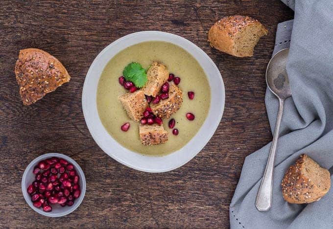 a table with a bowl of parnsip apple soup garnished with herbs, pomegranate seeds and bread. With a spoon and napkin on the side