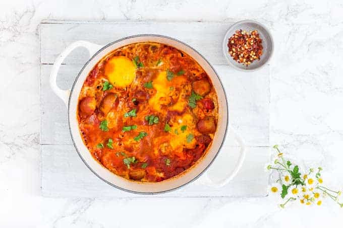 overhead view of easy shakshuka recipe in a large white pot on a white stone on a white countertop next to flowers