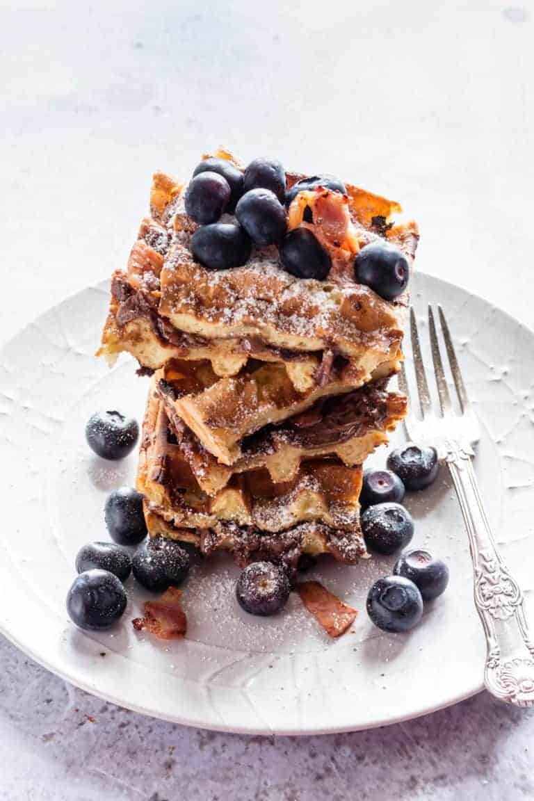stack of decadent nutella waffles on a white plate with berries, bacon and a fork