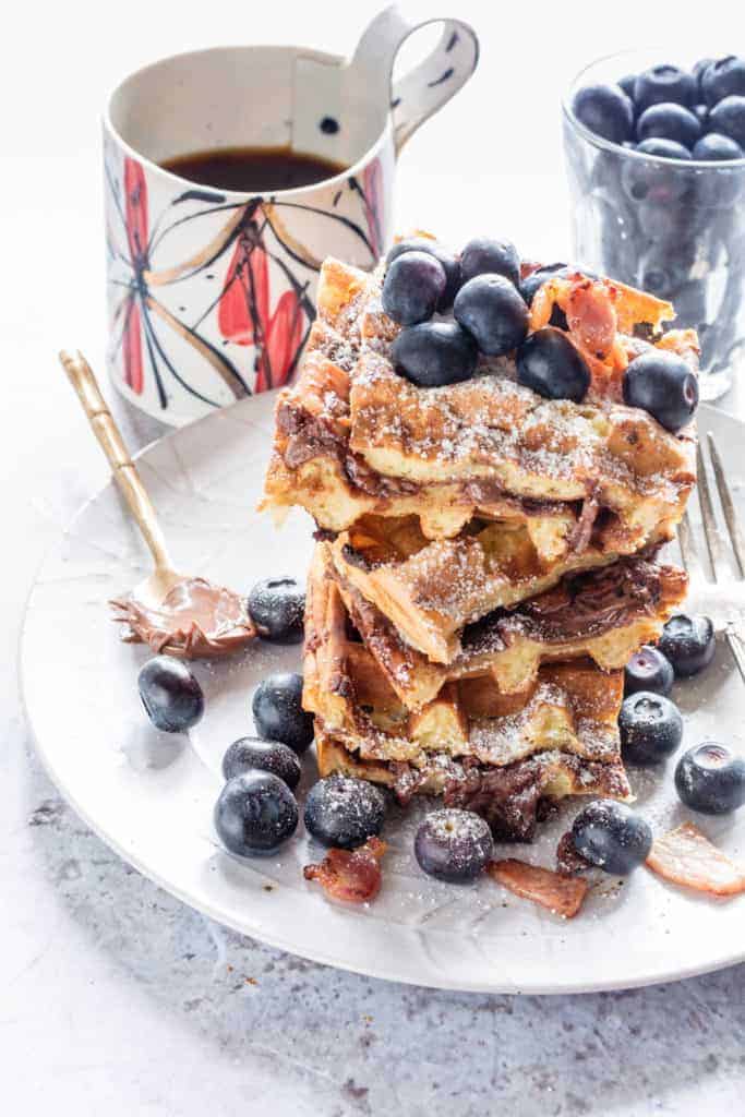 Crispy bacon Nutella  waffles on a plate surrounded by blueberries and bacon