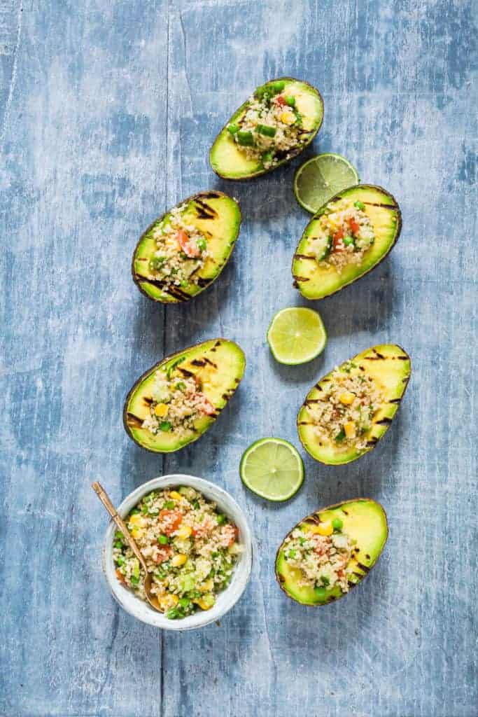 Six grilled avocado halves on a table stuffed with veggies and a bowl of veggie quinoa and teaspoon on the side. 