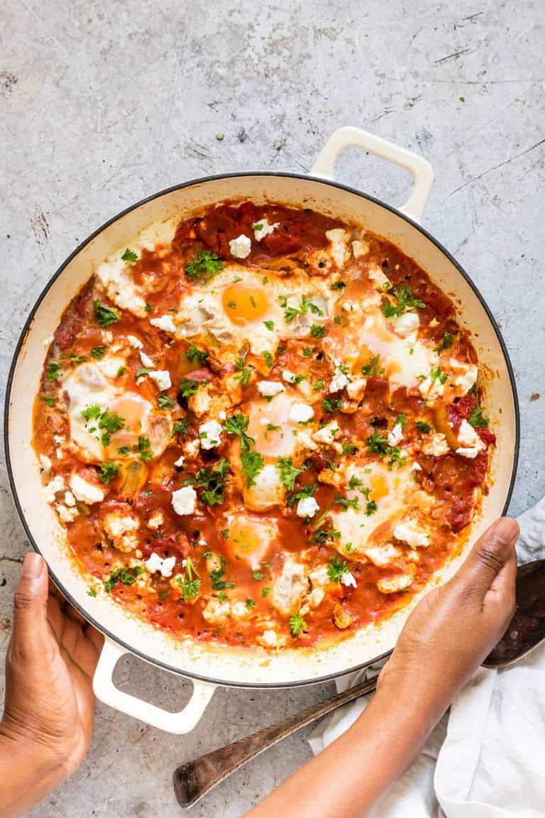 two hands holding a cast iron pot containing cooked shakshuka with feta