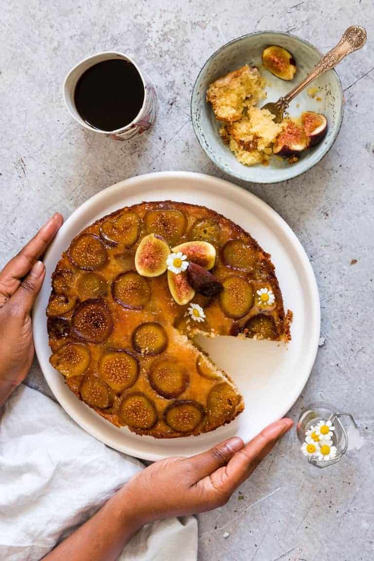 Hand holding an Upside Down Fresh Fig Cake, with a cup of coffee and a slice of the cake in a bowl