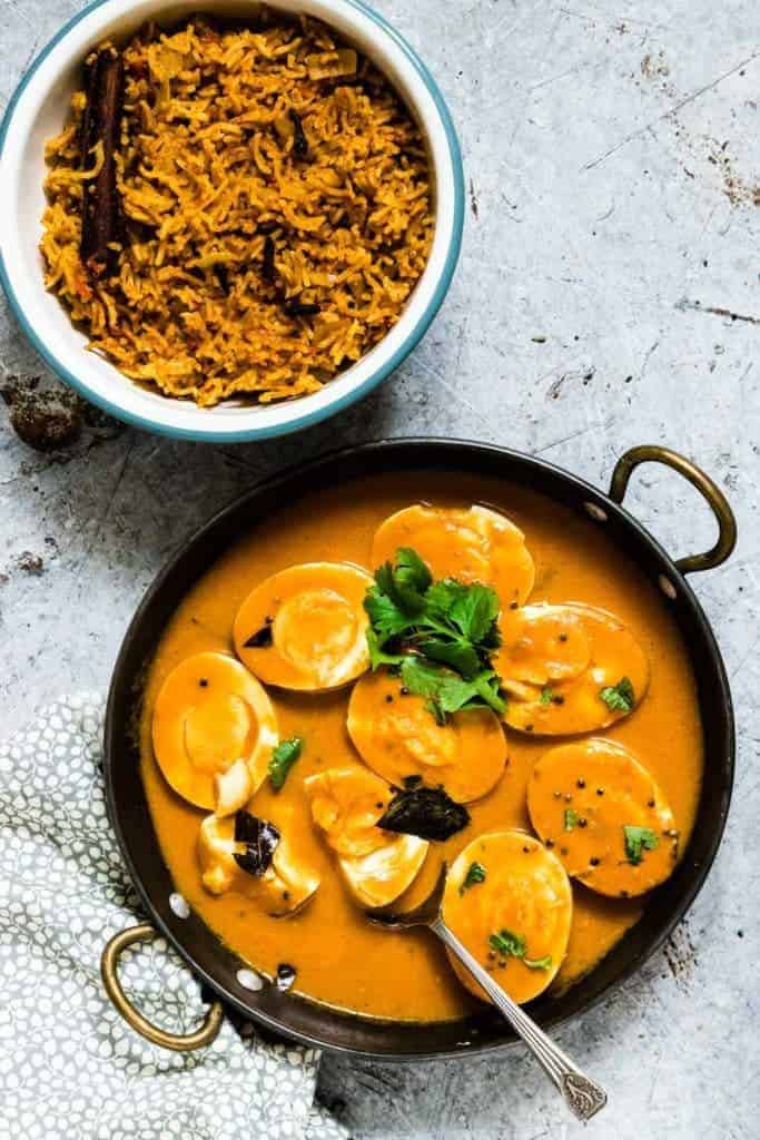 creamy Indian egg curry recipe in a pot with a spoon and some herbs and a bowl of tomato rice