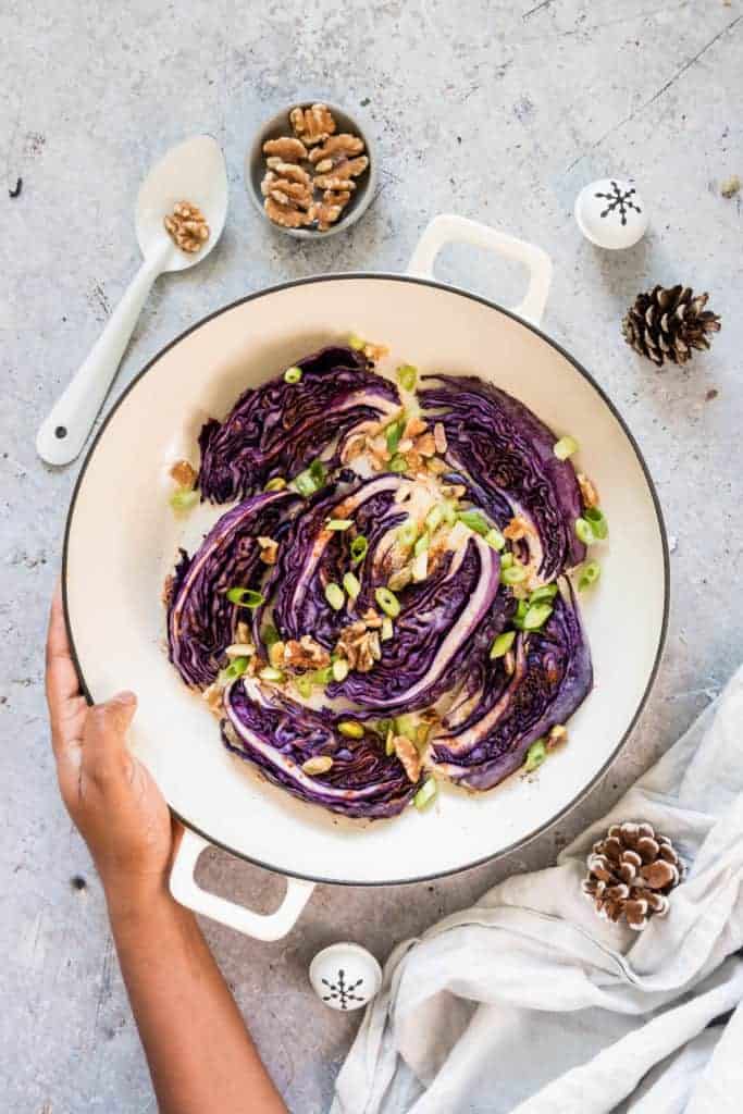 Roasted red cabbage in a pot with walnuts, spring onions, a spoon, a bowl of walnuts, cloth, pine cones and a hand