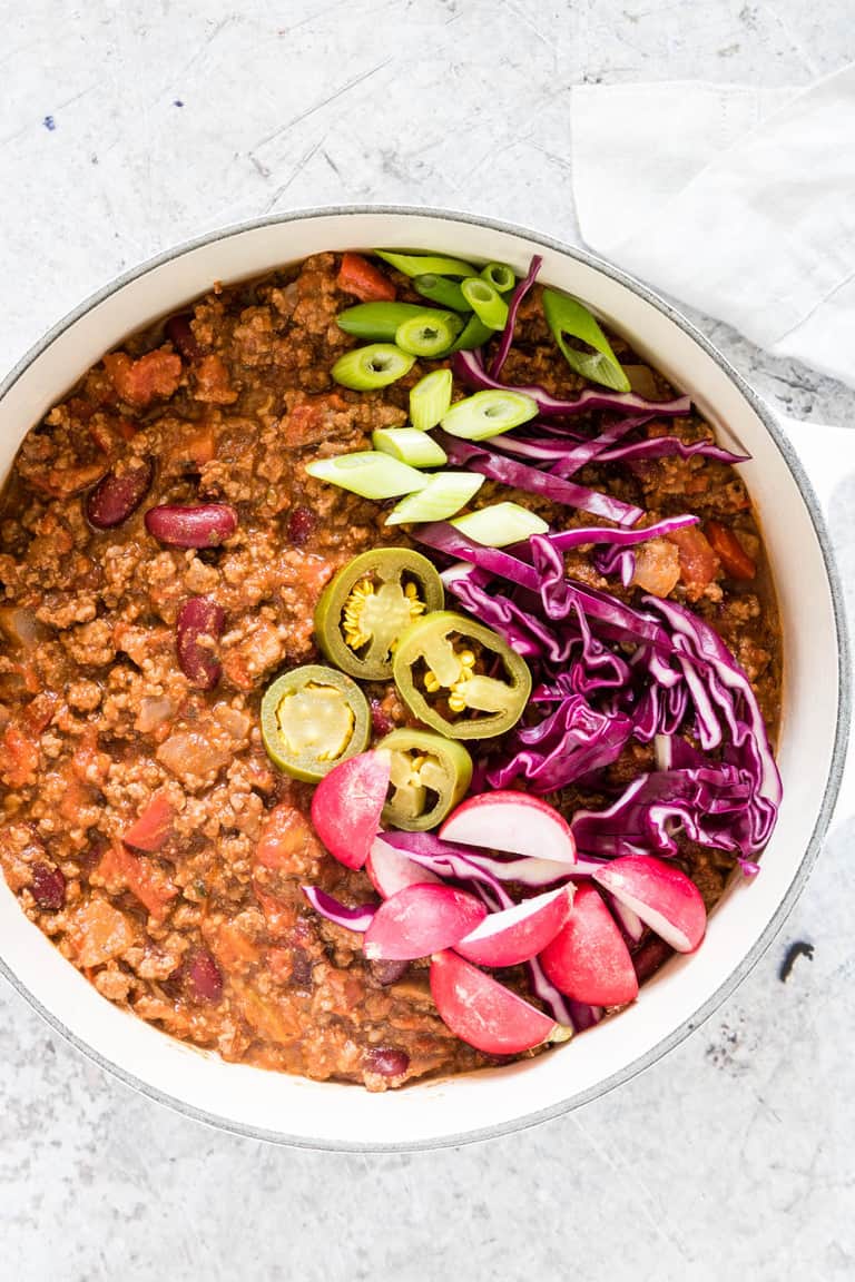 Overhead picture of cooked venison chilli in a white pot with spring onion, jalapenos, radishes and red cabbage as garnish