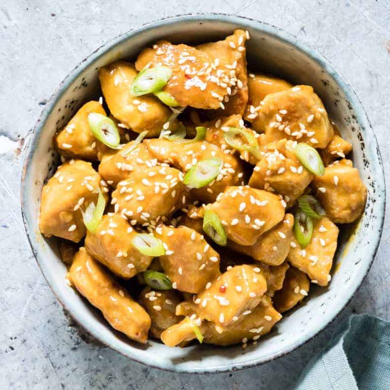 square closeup image of orange chicken in a blue bowl with sesame seeds on top