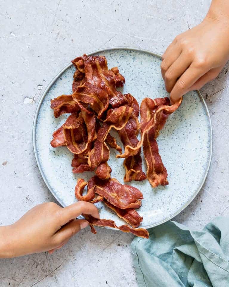 Two hands reaching for cooked strips of Crispy Air Fryer Bacon being served on a plate next to a blue cloth napkin