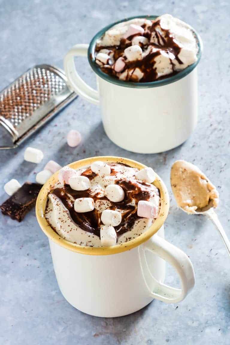 2 mugs of vegan hot chocolate with whipped cream, chocolate sauce, marshmallows