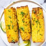 three pieces of Air Fryer Corn on the Cob topped with fresh herbs and served on a white plate with lime wedges