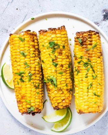 three pieces of Air Fryer Corn on the Cob topped with fresh herbs and served on a white plate with lime wedges