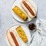 two white plates containing Air Fryer Hot Dog and Air Fryer Corn on the Cob, served with a small condiment dish and a cloth napkin