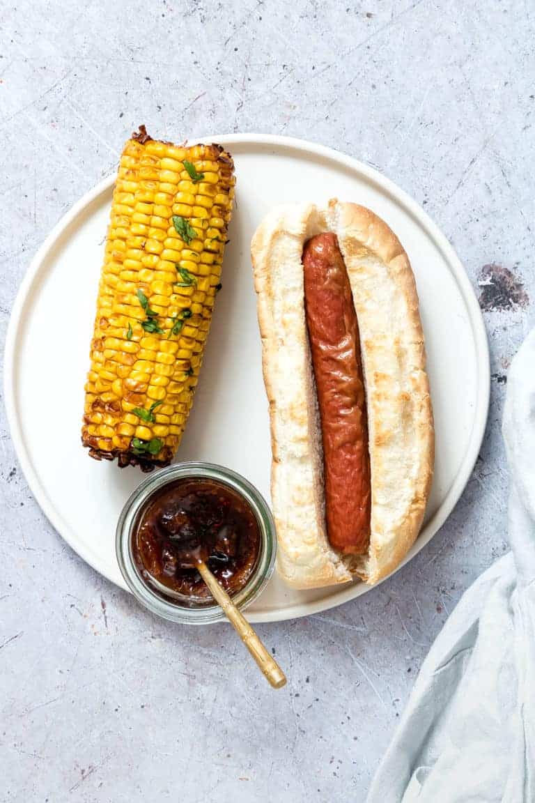 one hot dog served with an ear of air fryer corn on the cob and a small glass condiment dish 
