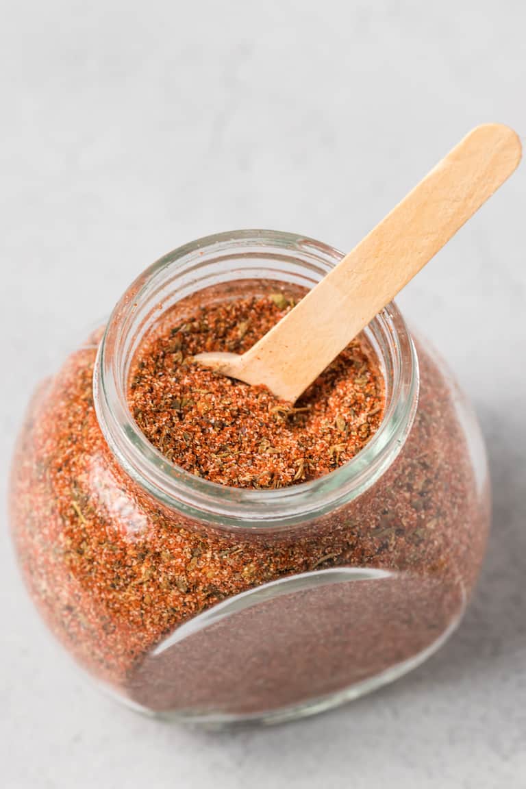 a storage jar filled with creole seasoning with a wooden measuring spoon inside