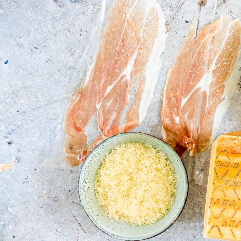 table with ingredients for cheesy quinoa porridge including two thin slices of prosciutto and bowl of grated cheese