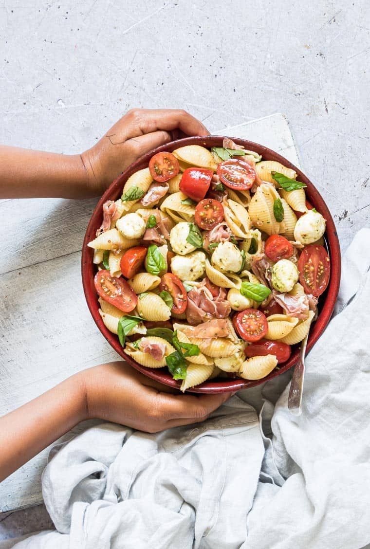 two hands setting out a red bowl filled with caprese pasta salad