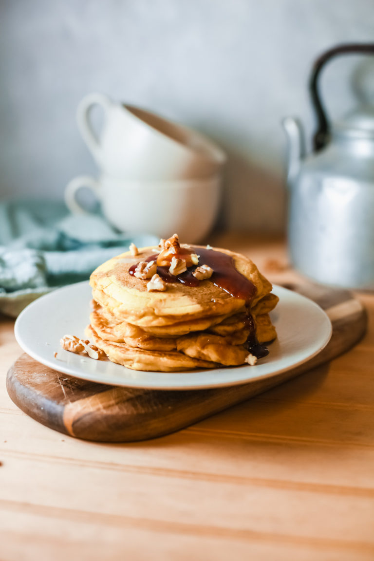 a stack of pumpkin pancakes served on a white plate and topped with butter, syrup and nuts