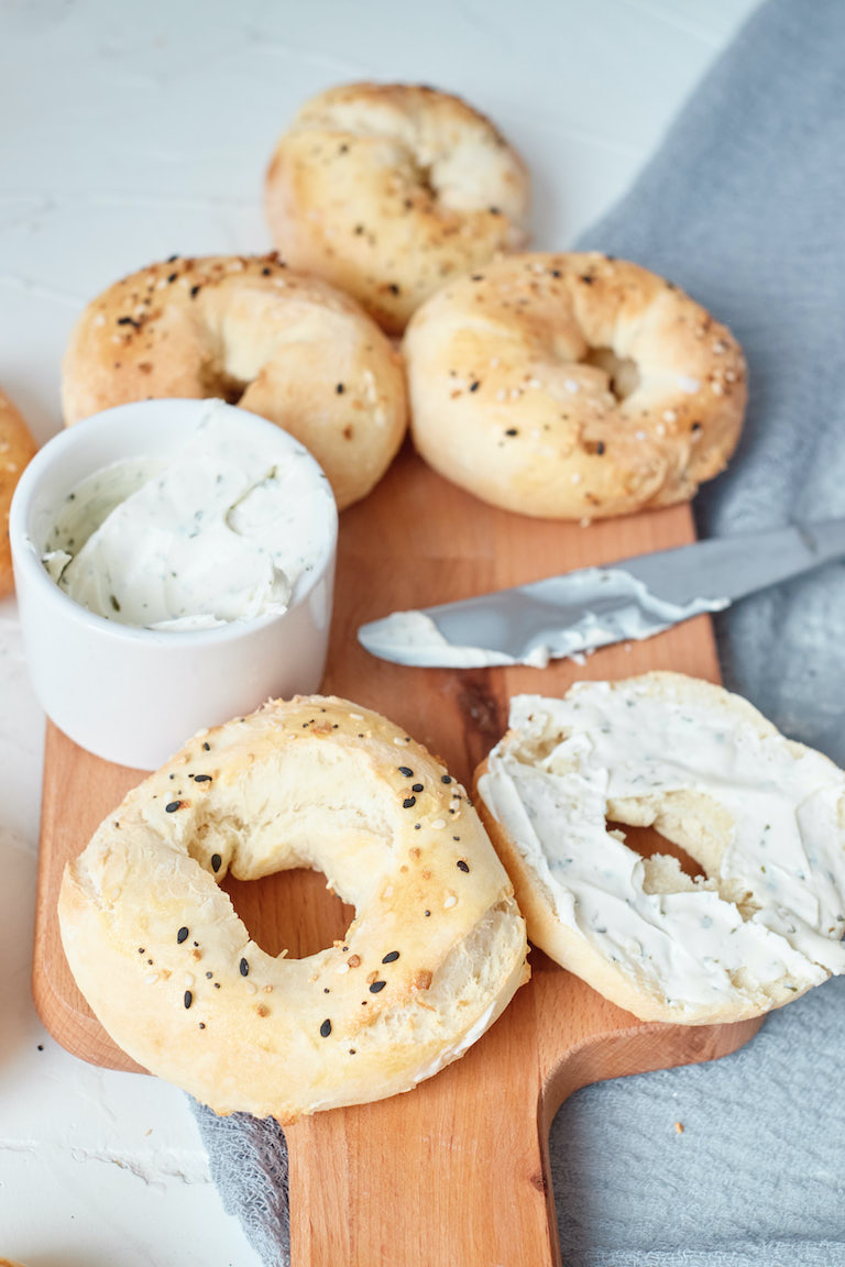 four everything bagels on a cutting board with one sliced in half and spread with cream cheese
