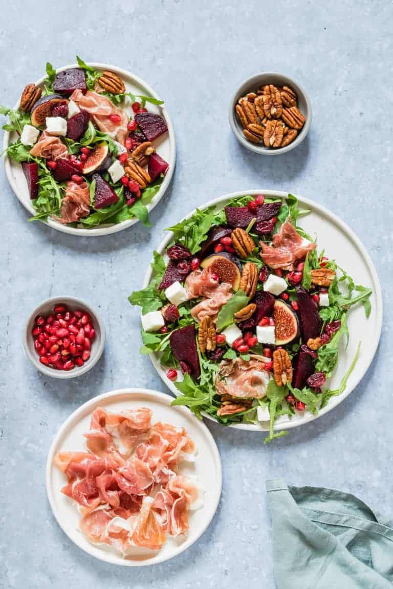 Parma Ham and Beet Salad served on white plates placed next to a small bowl of pecans, a small dish of pomegranate seeds, and a small plate of Parma Ham 