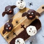 Halloween cupcakes decorated to look like bats and skulls set on a wooden cutting board painted with a graveyard scene along with plastic toy spiders