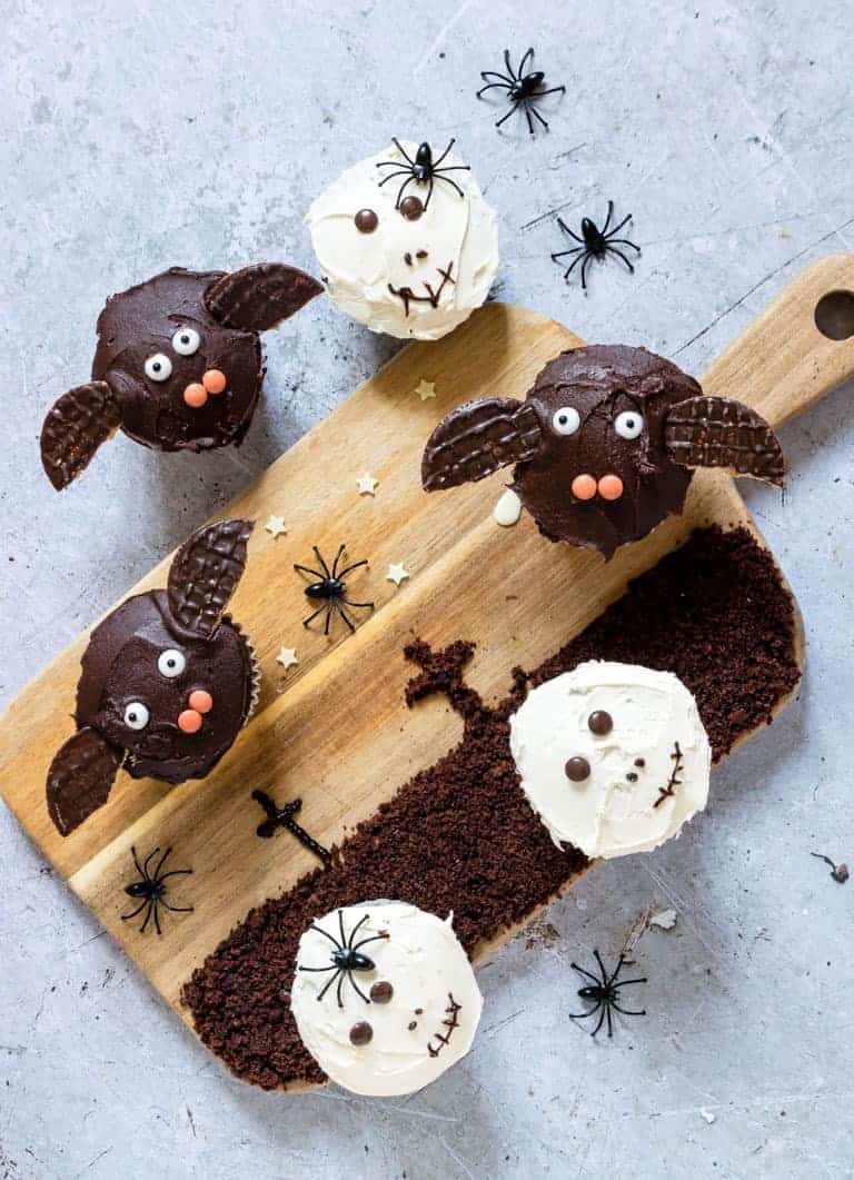 Halloween cupcakes decorated to look like bats and skulls set on a wooden cutting board painted with a graveyard scene along with plastic toy spiders