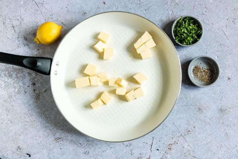 cubed butter added to a light-coloured saucepan with lemon and fresh herbs, the other ingredients for Lemon Butter Sauce, placed nearby