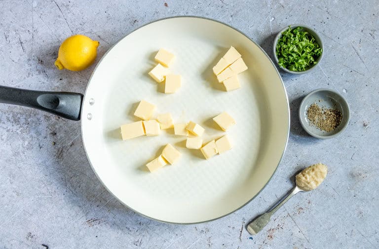 Cubed butter inside a light-coloured saucepan with the remaining ingredients for Lemon Garlic Butter Sauce, lemon, garlic puree, fresh herbs and black pepper, sitting next to the saucepan