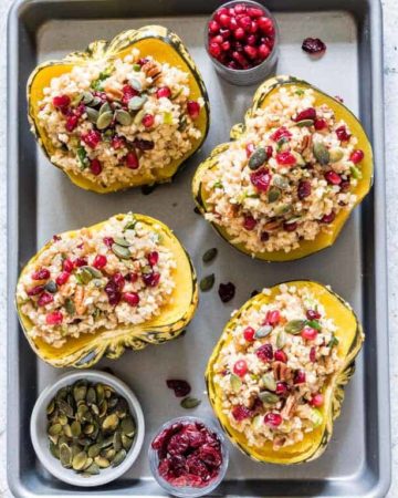 Four servings of Instant Pot Stuffed Squash on a baking sheet alongside small bowls of pumpkin seeds, cranberries, and pomegranate seeds