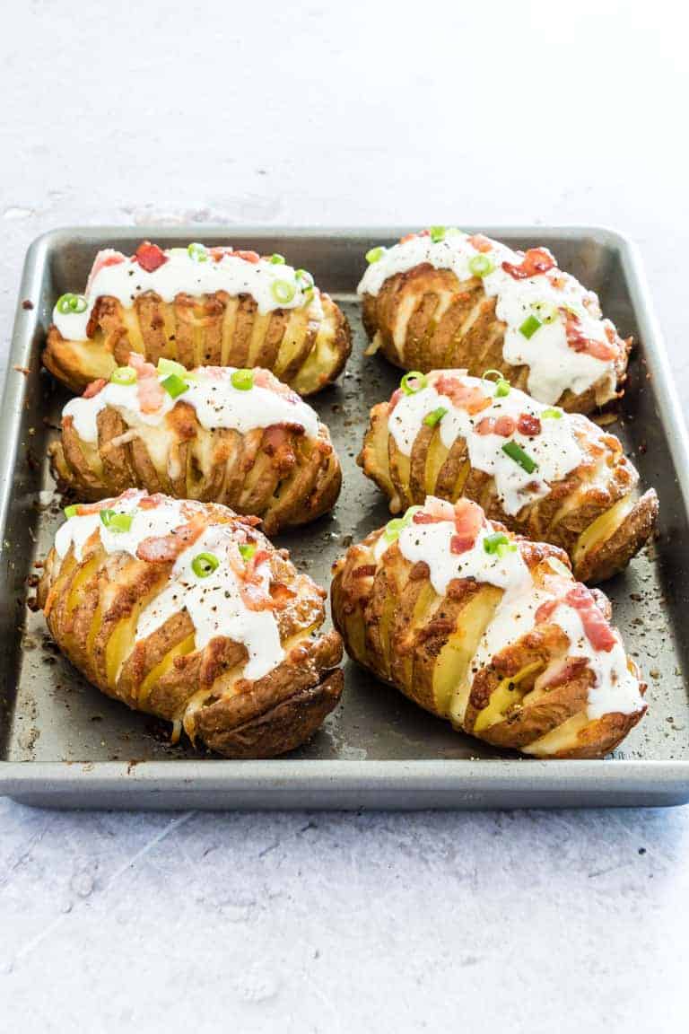 Six Loaded Hasselback Potatoes topped with sour cream, spring onions and bacon lined up on a baking sheet and ready for serving