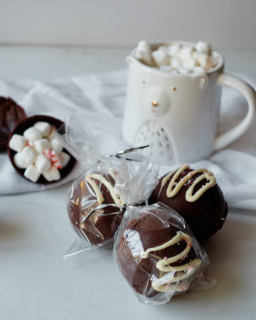 three chocolate bombs packages as edible gift ands placed in front of a mug of hot chocolate