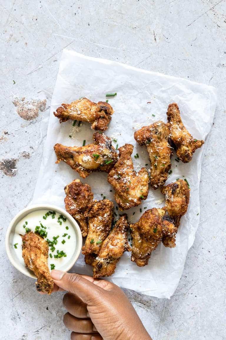 A hand dipping one of the Air Fryer Chicken Wings into ranch sauce next to the rest of the chicken wings that are set on white parchment paper 