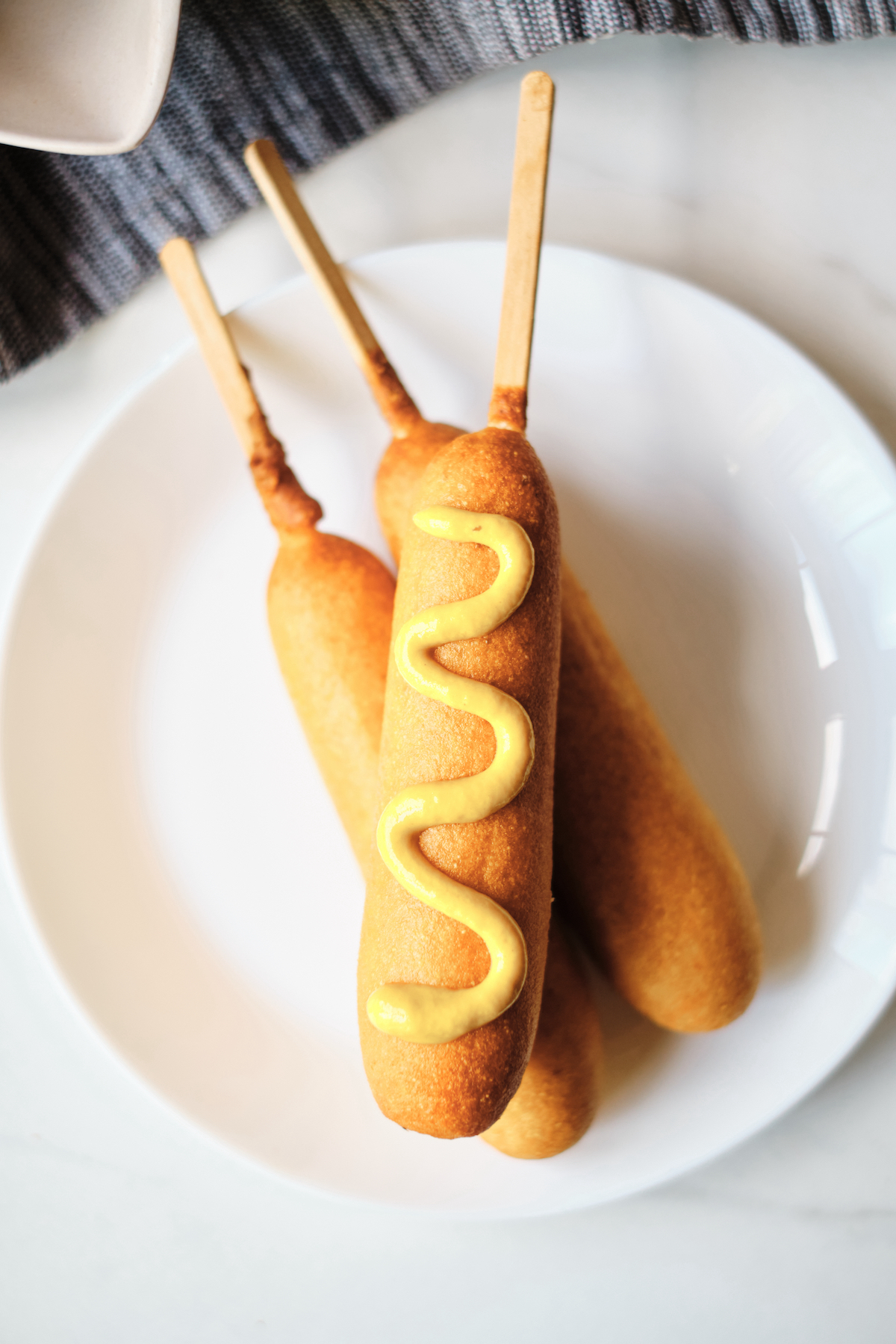 three frozen corn dogs that have been cooked in air fryer on a white plate and topped with mustard