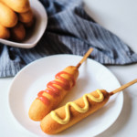 two cooked frozen corn dogs on a white plate one topped with ketchup and one topped with mustard