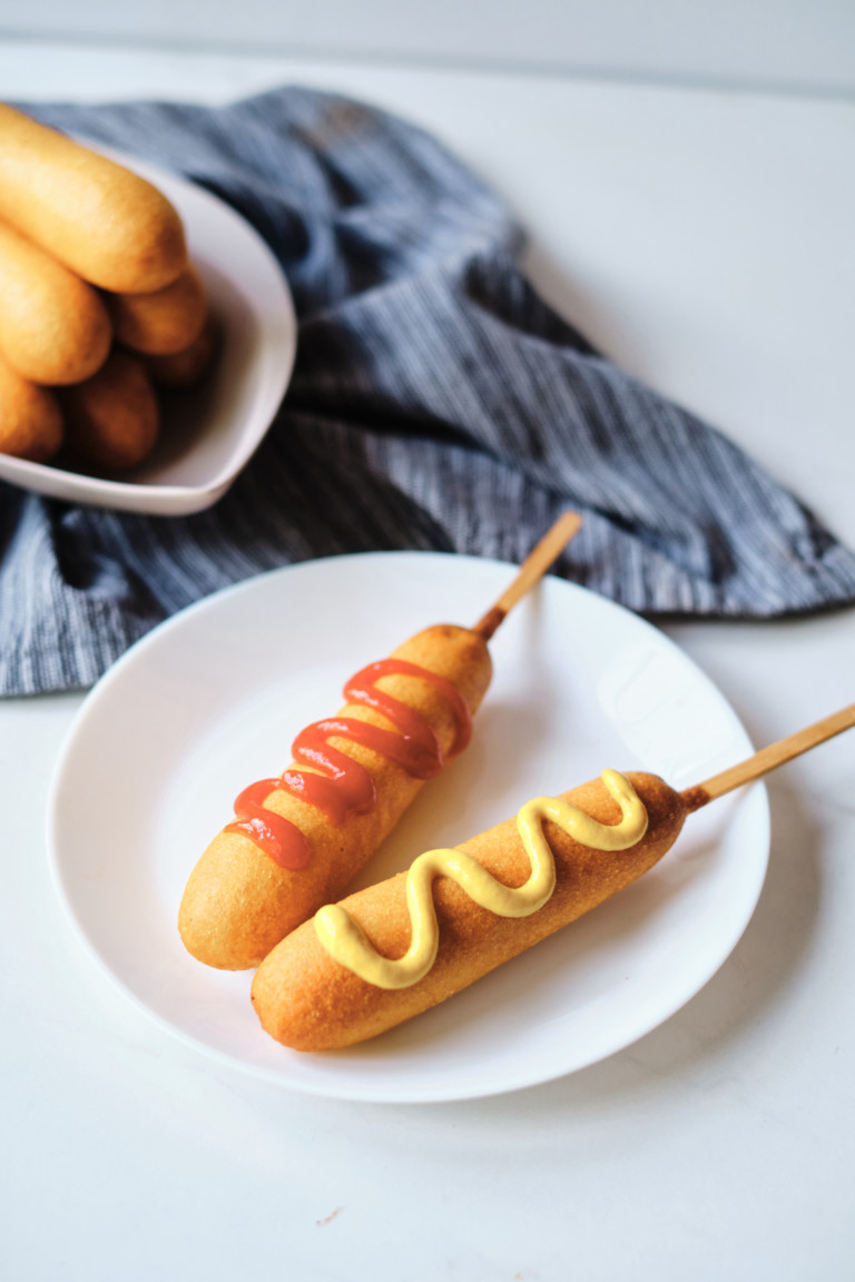 two cooked frozen corn dogs on a white plate one topped with ketchup and one topped with mustard