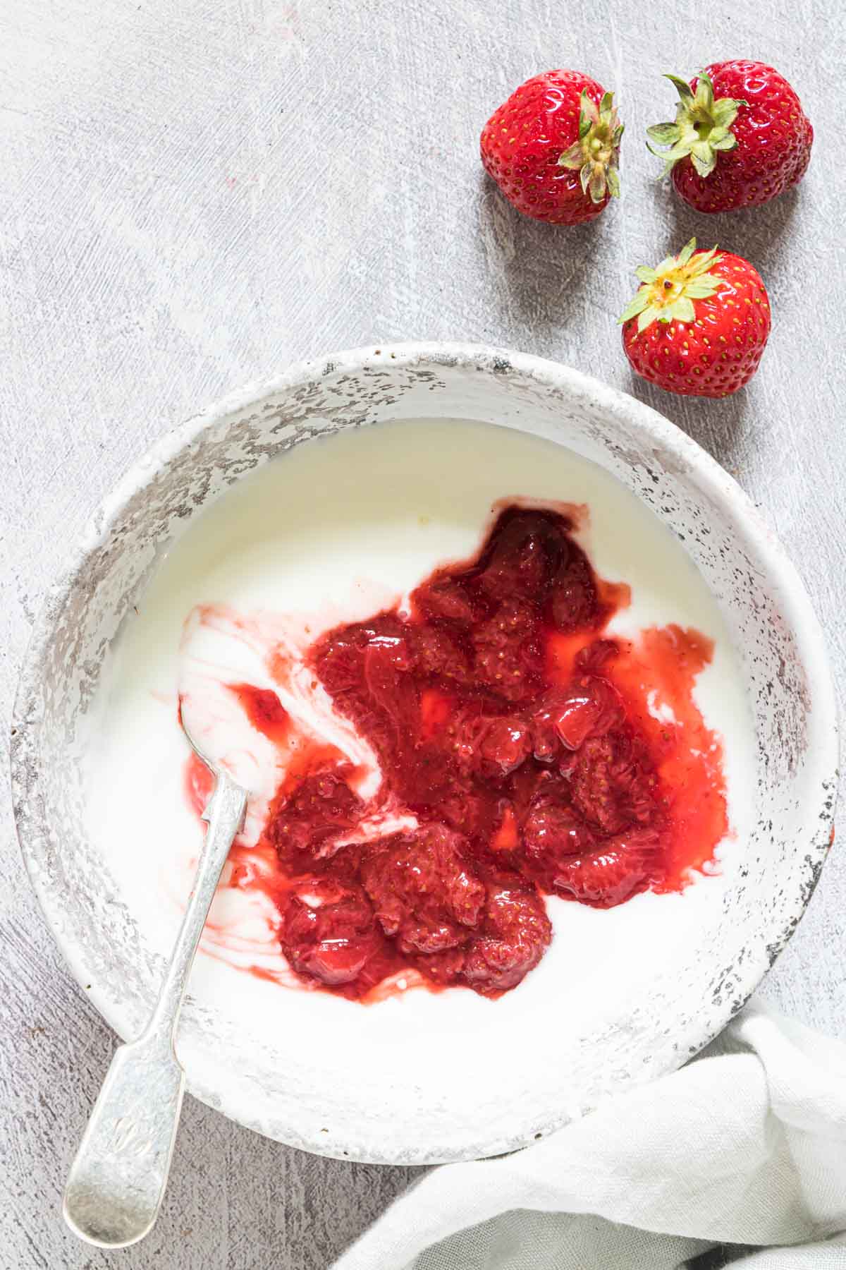 a bowl of yogurt with homemade strawberry sauce on a table with a spoon added in and 3 strawberries