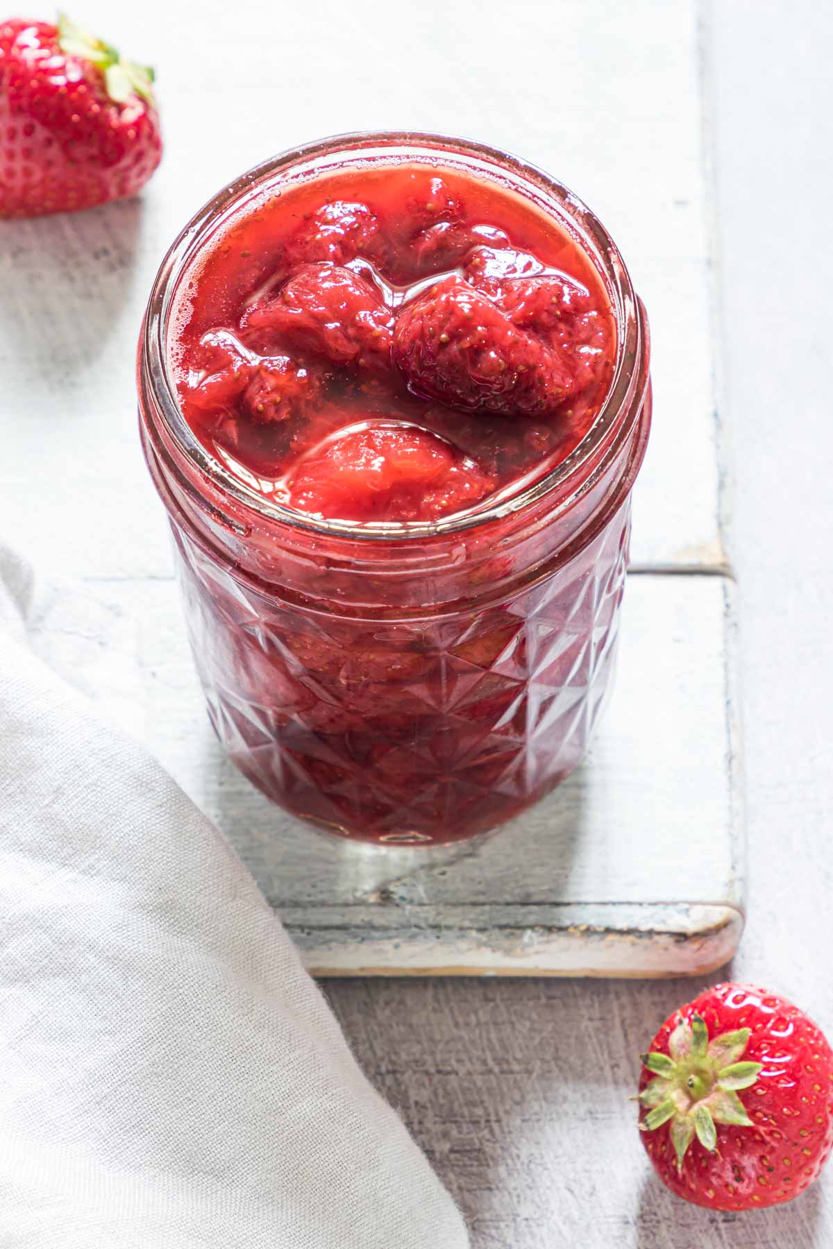 a jar of homemade strawberry compote on a chopping board with a couple of strawberries