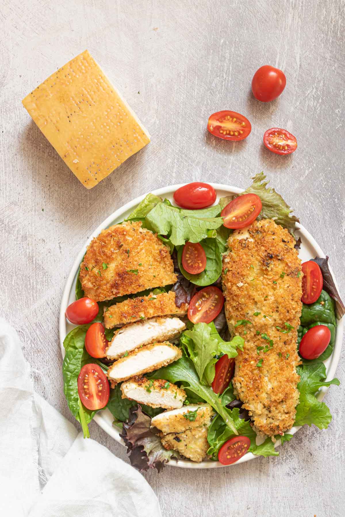 a serving of parmesan crusted chicken breast with a green salad on a white plate next to a wedge of parmigiano reggiano cheese