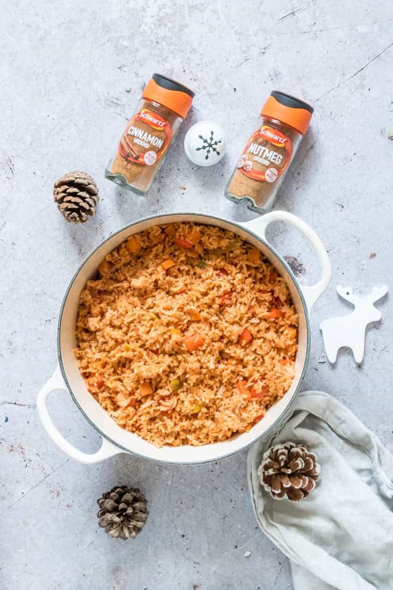 Cinnamon Spiced Jollof Rice in a large white pot next to a jar of cinnamon, a jar of nutmeg, pinecones and white christmas ornaments