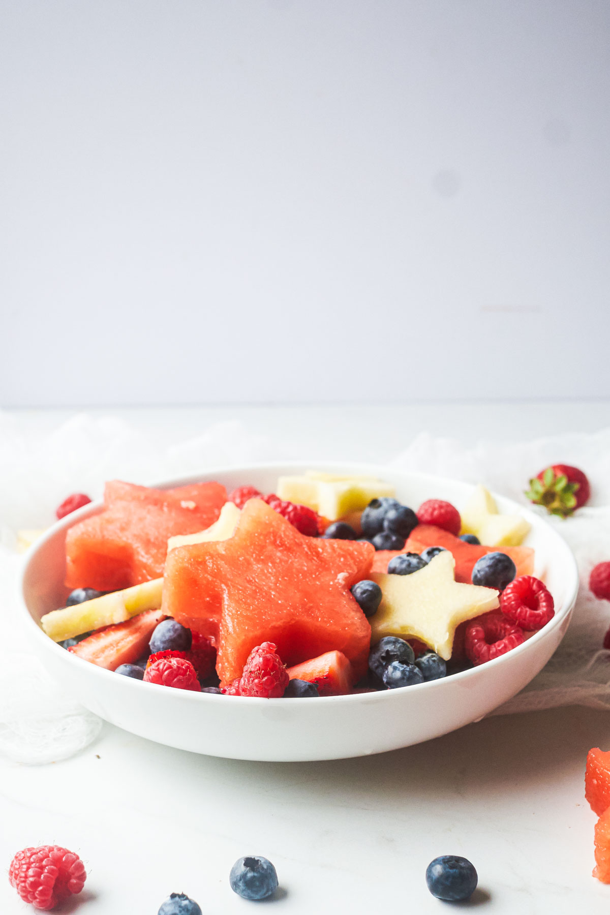 side view of a red white and blue fruit salad on a white tables, with some fruit dotted about.