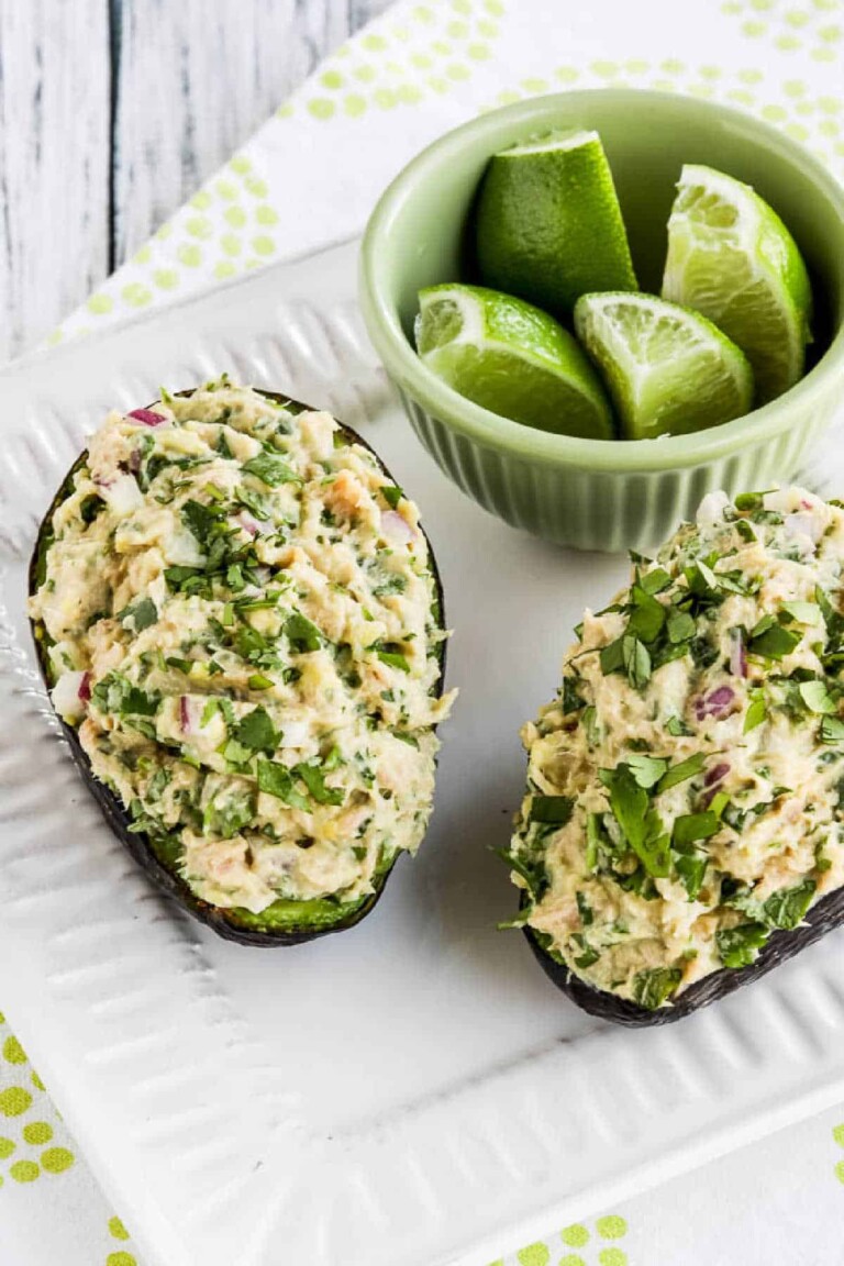 Two Tuna Stuffed Avocados on a white plate with limes in a green bowl.