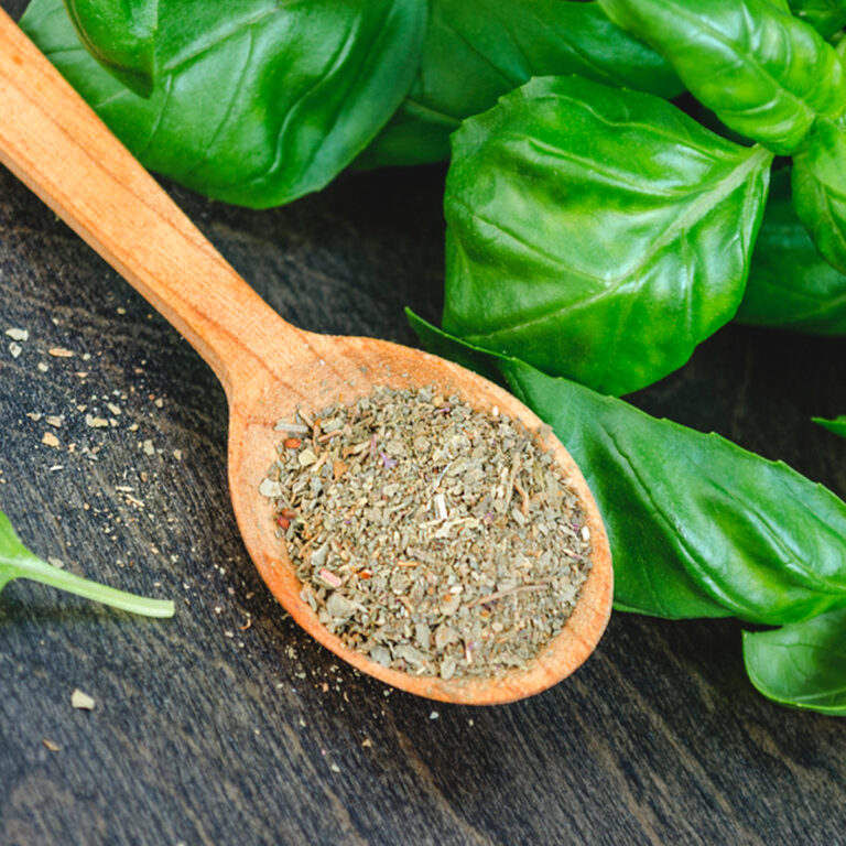 A wooden spoon full of dried basil with fresh basil leaves surrounding it.
