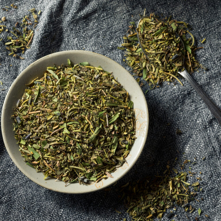 A ceramic bowl of herbs de Provence on a blue cloth with a spoonful of herbs beside it.
