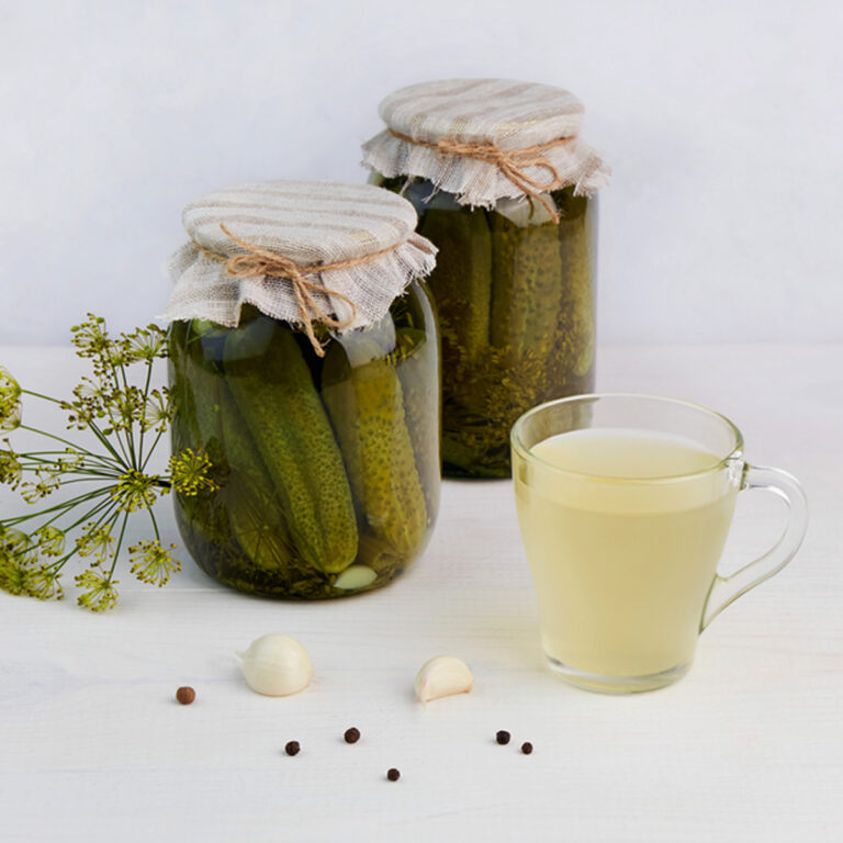 Two Jars of pickles with a glass of pickle juice in the foreground and a sprig of dill flowers beside them.