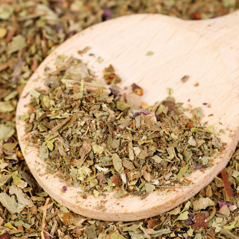 Close up of wooden spoon with dried flakes of herbs on it, laying in a bed of herbs.
