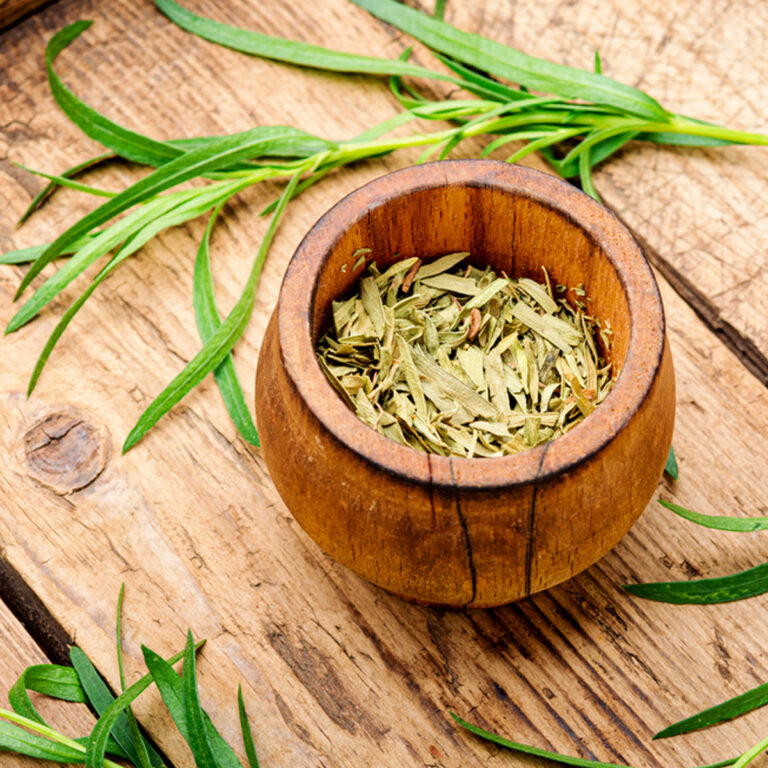 A small wooden bowl of dried tarragon on a wooden tabletop with a sprig of fresh tarragon behind it.
