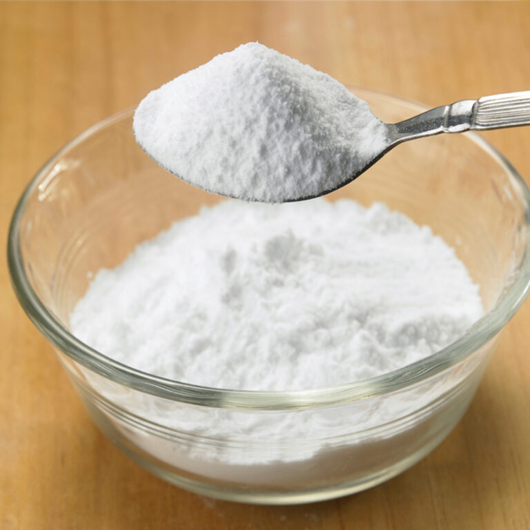 A spoon taking a scoop of baking powder out of a glass bowl