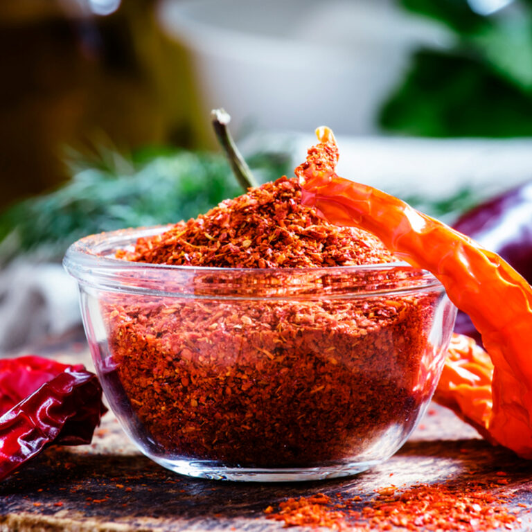 A glass bowl of ground cayenne pepper with whole dried peppers beside it.
